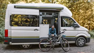 Mann sitzt auf der Treppe eines weißen CamperBoys Camper, der auf einem Campingplatz in Holland steht und lächelt in die Kamera, vor ihm steht sein Fahrrad